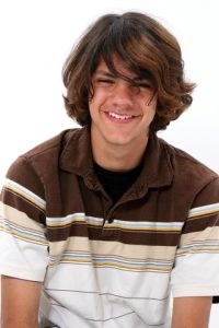 A smiling teenage boy in a brown, tan and white striped shirt with shaggy brown hair.