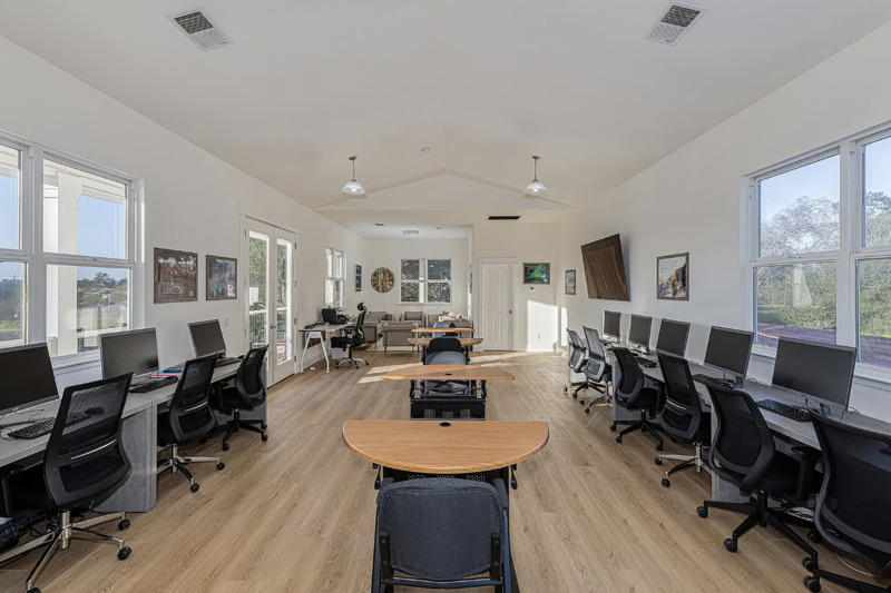 A rectangular room with a row of desks down the middle of it and desks with multiple computers and chairs down each of the outside walls of the room.