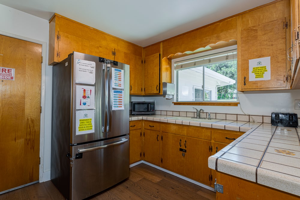 A kitchen with white tile countertops, light wood cabinets, wood floors, and a microwave and toaster on the counter.