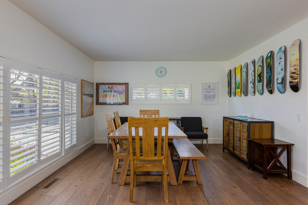 A room with a wooden dining table, a cabinet, two gray chairs, skateboard decks on one wall, and windows.