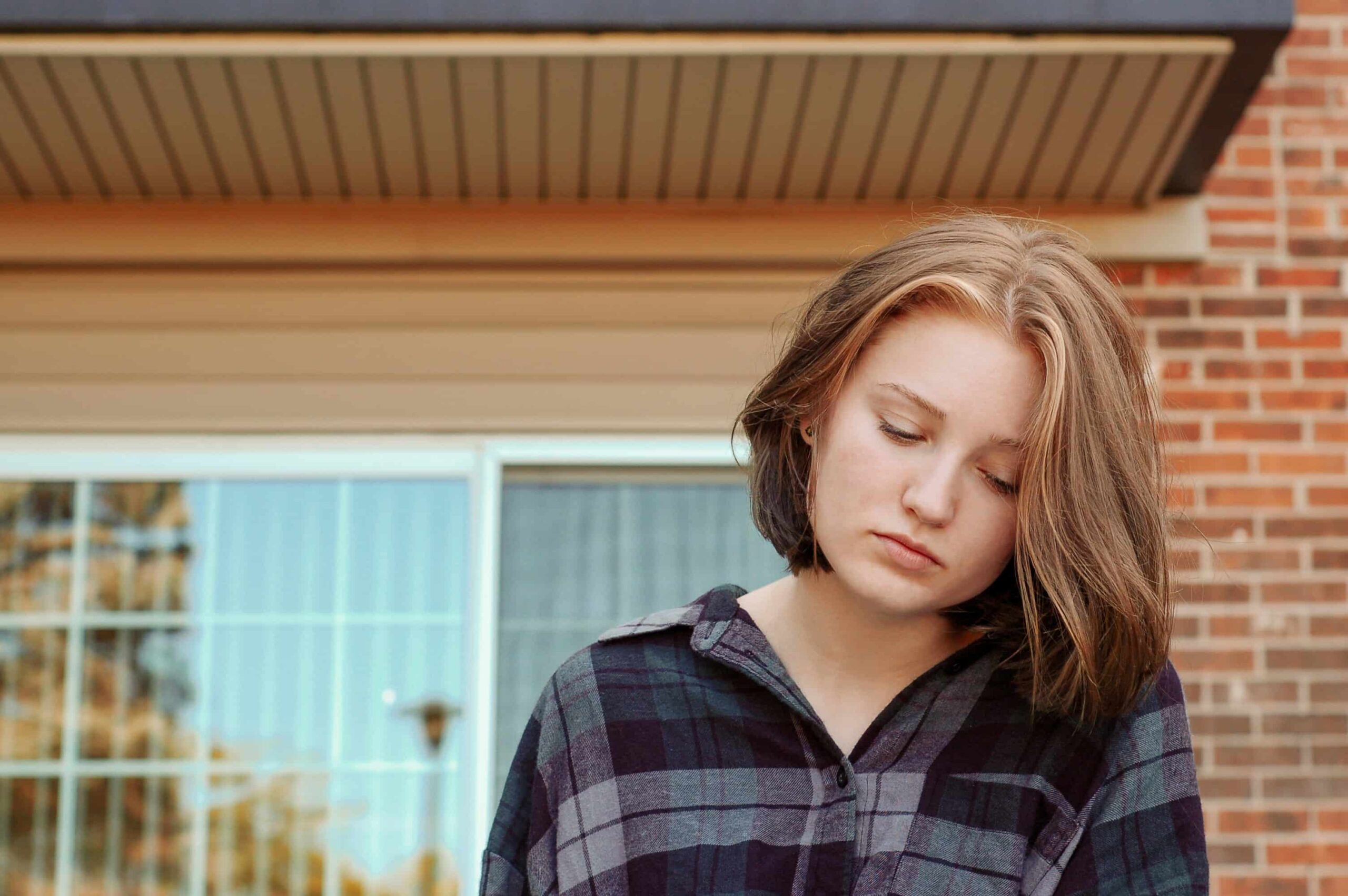 Young woman standing outdoors, looking down, wearing a plaid shirt in front of a brick building.
