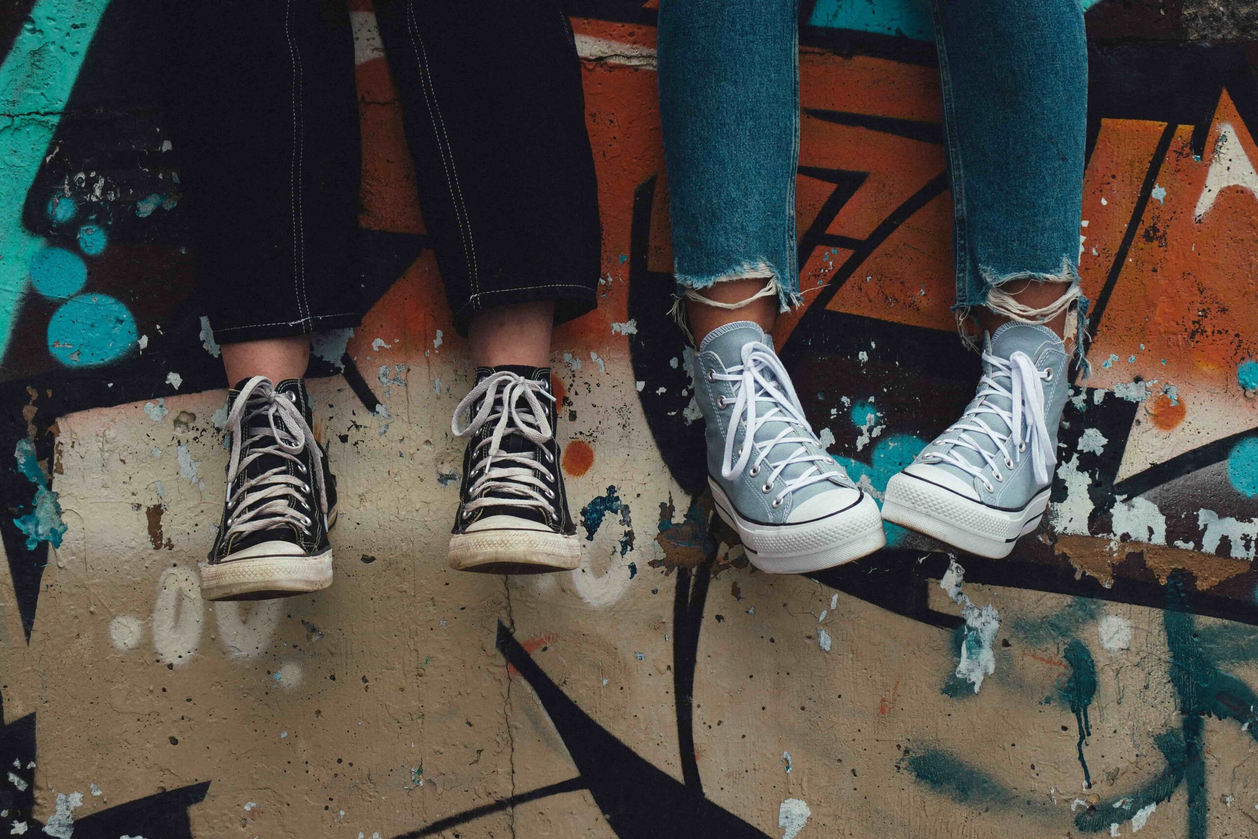 Two people sitting side by side with their legs dangling, wearing Converse sneakers in front of a colorful graffiti wall.