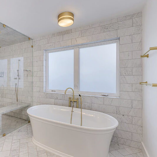 A bright bathroom with a glass shower and standalone porcelain tub. The room has gold fixtures.
