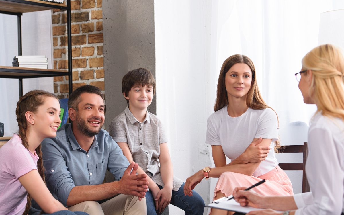 A diverse family sits together in a bright office while engaging in a supportive therapy session with a clinician.
