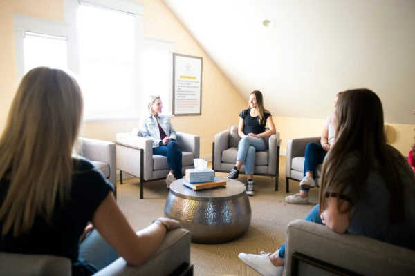 A female therapist leads a group session with four teenage girls sitting in a bright, comfortable clinical room.