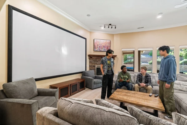 Four teenage boys talk and relax in a comfortable residential media room featuring a large projector screen and gray sofas.