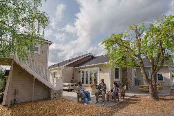 Three teenage boys sit in wooden chairs on a sunny outdoor patio behind a residential campus with trees and a staircase.