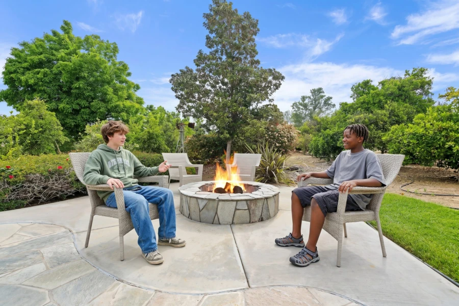 Two teenage boys sit in comfortable chairs by a stone fire pit on a sunny outdoor patio surrounded by green trees.