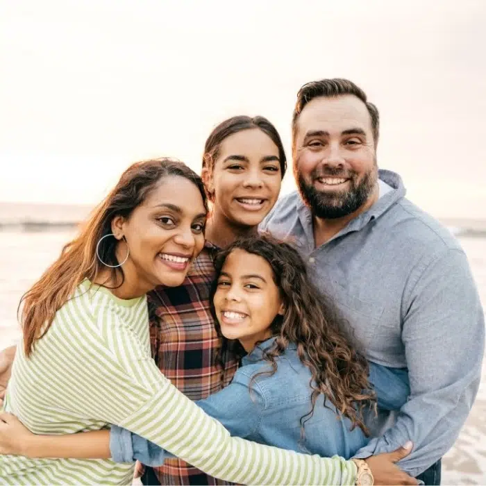 A smiling family of four, including two parents and two children, share a warm hug outdoors near the beach.
