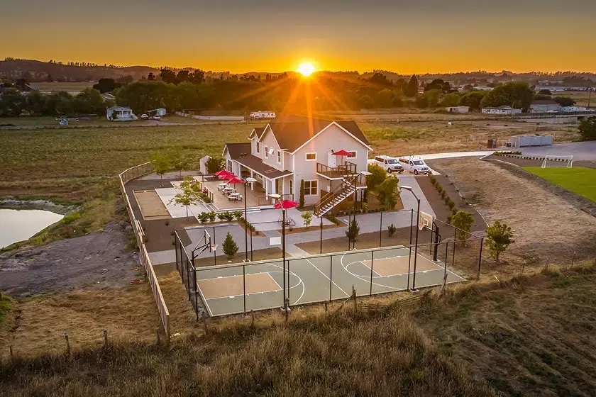 An aerial sunset view of a large residential facility featuring a basketball court, patio, and green hills.
